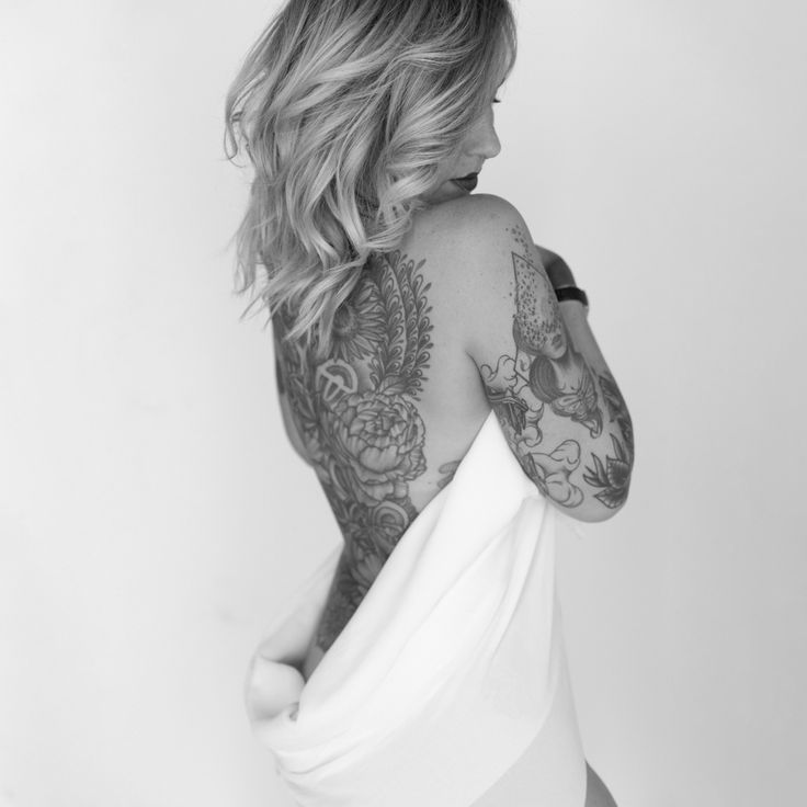 a black and white, closeup portrait of a woman's back, showcasing an intricate floral tattoo that extends from her lower back up to her shoulders. her blonde, wavy hair partially covers one shoulder.  she is partially draped in a white cloth, creating a sense of modesty while still highlighting her tattoos. the overall tone of the image is sensual, artistic, and elegant, suggesting themes of body positivity and selfacceptance. the background is a simple, minimalist white.
