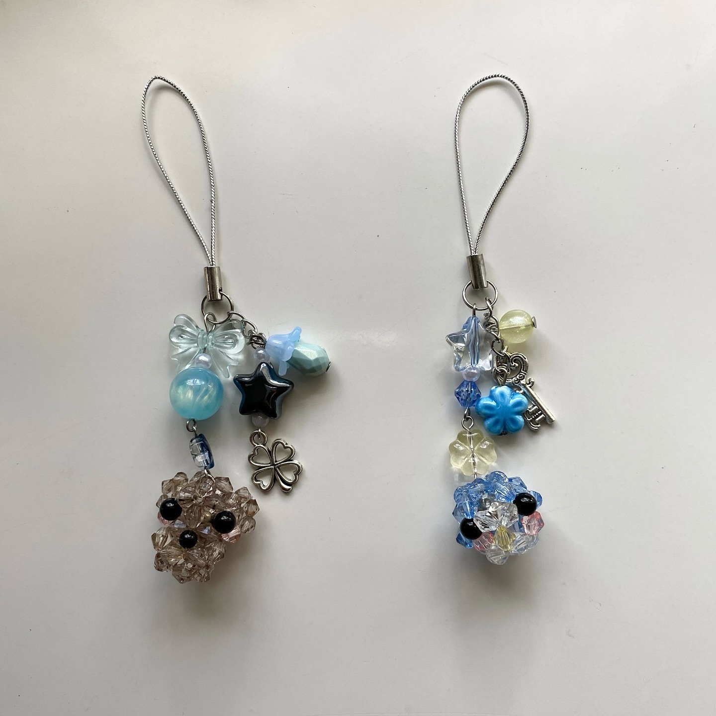 two beaded charms are lying side by side on a white surface. the charm on the left features a beaded dog with a tan tint, a black star, a fourleaf clover, a light blue bow, and blue beads. the charm on the right also features a beaded dog with a blue tint, as well as a star, a flower, a key, and a mix of blue and yellow beads. both charms have silver phone straps attached.