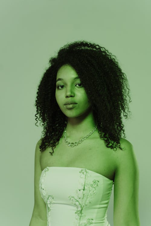 closeup portrait of a young africanamerican woman with curly hair, wearing a white strapless dress and a delicate necklace. the image is tinted green, giving it a stylized and slightly mysterious look.  she is looking directly at the camera with a serious expression. the background is a plain, light green.