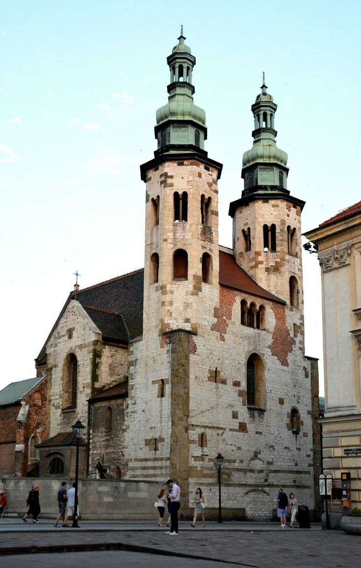 the image shows the church of st. andrew in krakow, poland.  it's a prominent medieval church, easily recognizable by its twin romanesque towers capped with green roofs. the church's exterior is a mix of stone and brick, showcasing varying shades and textures.  the photograph captures the church from a slightly low angle, emphasizing its height and the intricate details of its facade. several people are visible in the foreground, strolling on a cobblestone street, offering a sense of scale and indicating the church's location within a bustling urban environment. the sky is a pale, almost clear blue, suggesting daytime. a portion of another building is visible to the right, providing context to the church's urban setting.