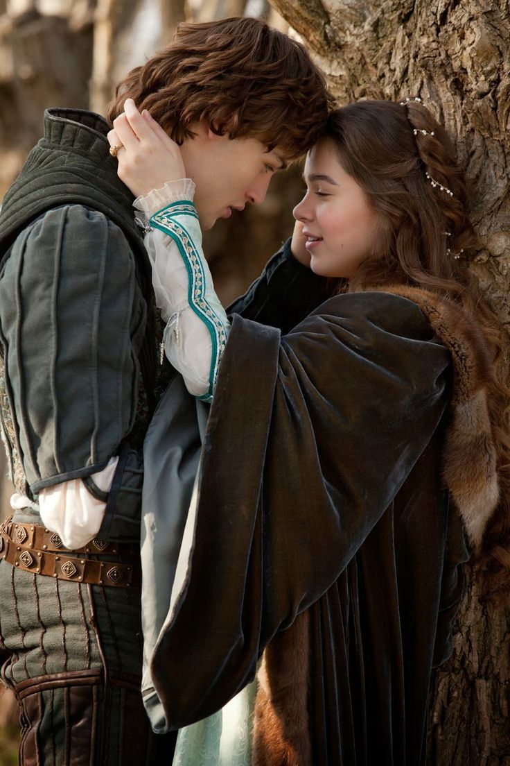 a young couple, dressed in medieval clothing, embrace and look lovingly at each other in a forest.  the man is wearing a dark green jacket, a white shirt, and brown pants. the woman is wearing a dark green velvet robe with a brown fur trim. the couple is standing under a large tree, with the woman's hand resting on the man's cheek.  the man is looking at the woman with a warm, loving gaze, while the woman is looking at the man with a shy smile.