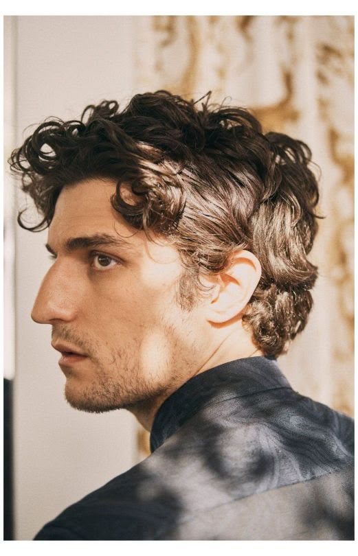 a profile portrait of a man with curly dark hair and light facial hair, wearing a dark shirt. he's looking to the left.