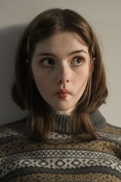 a young woman with brown hair wearing a knitted sweater. the lighting is soft and the perspective is at eye level.  she is making a funny face, pursing her lips together in a slightly pouty expression, while her eyes are wide open.  her hair is styled with a middle part and bobby pins holding it back on each side.