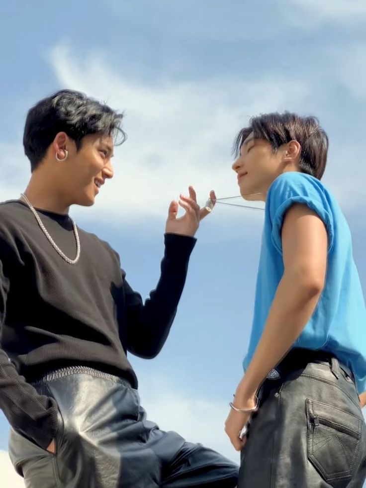 the image features two men outdoors looking at each other. one is wearing a black shirt and leather pants and the other is wearing a blue shirt and leather pants. the man in the black shirt is smiling and holding a chain necklace while the man in the blue shirt looks at him lovingly. the background is a blue sky with clouds. the image suggests a romantic or close friendship between the two men.