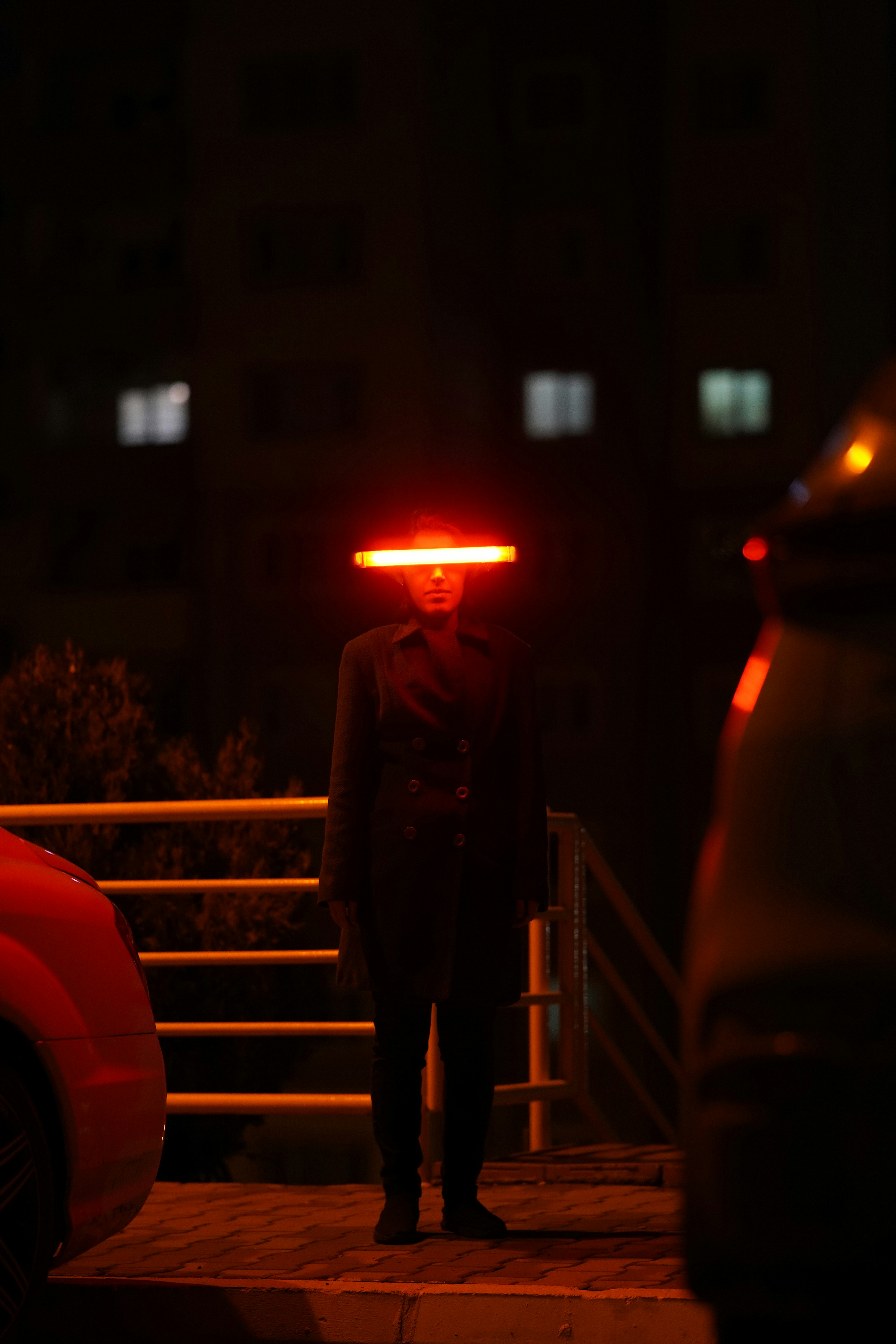 woman stands on a city street at night. she is wearing a black coat and a futuristic headband with a bright orange neon light emanating from it. the background is dark, with buildings and cars barely visible. the image has a cyberpunk and futuristic aesthetic.