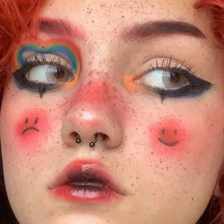 a closeup portrait of a woman's face with colorful makeup. she has freckles and is wearing a nose ring. her eyeshadow is a vibrant mix of colors, and she has black eyeliner. there's a sad face drawn on one cheek and a happy face on the other. her lips are glossy, and her hair is red.