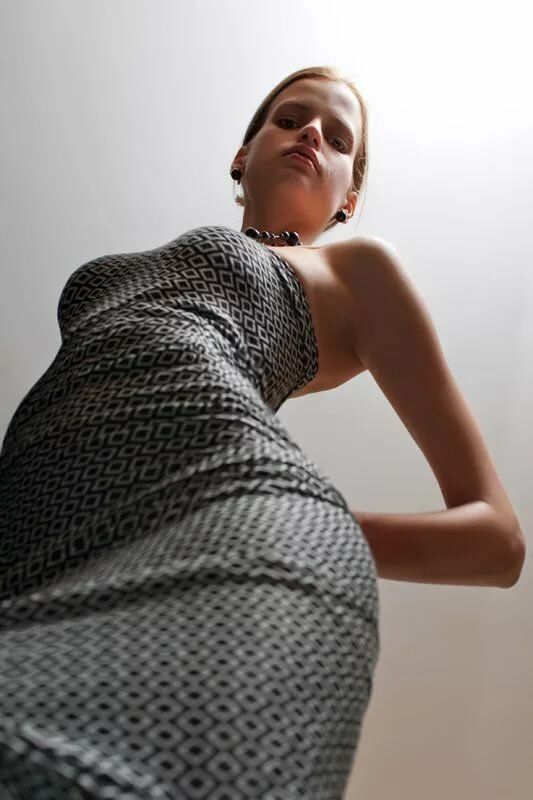 a woman in a geometricpatterned dress is standing in a room. the perspective is from a low angle, looking up at the woman. the lighting is natural, coming from a window or skylight above.