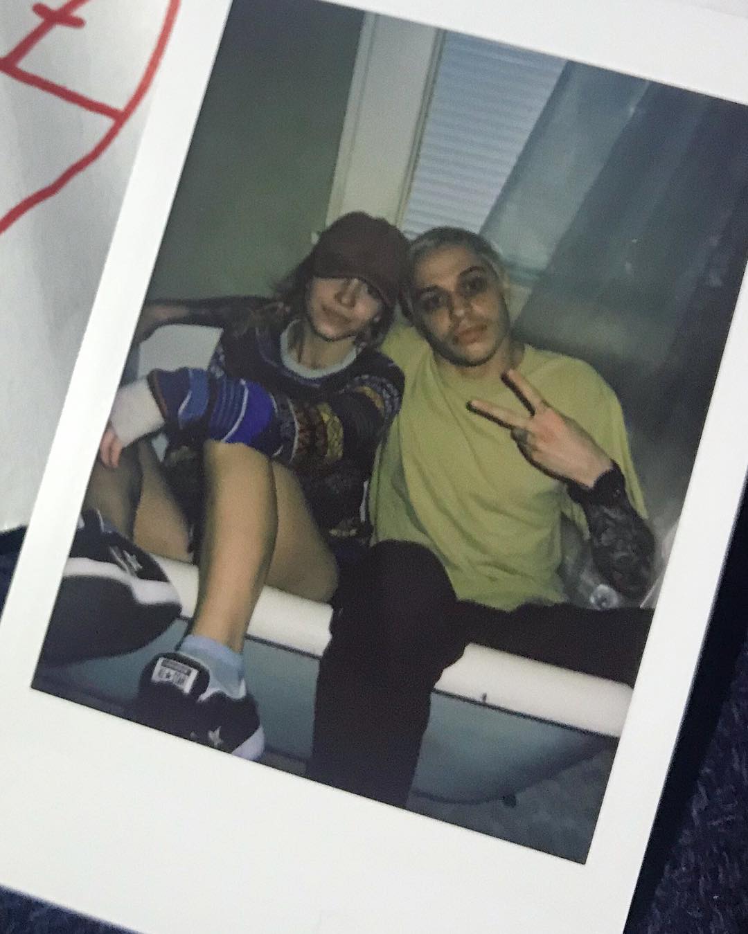 a polaroid photo shows a young couple sitting together in a bathtub. the girl is wearing a hat and a patterned sweater, and the boy is wearing a yellow tshirt and dark pants.  the boy makes a peace sign with his hand. they appear to be comfortable and relaxed, capturing a candid moment. the photo has a slightly grainy and faded quality consistent with instant film photography.