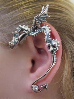 a closeup shot shows a woman's ear adorned with a silver dragon ear cuff. the earring wraps around the ear, featuring intricate details of a dragon's head, wings, and body. a small gemstone is located at the bottom of the cuff. the image captures the fantasyinspired and fashionable nature of the jewelry.