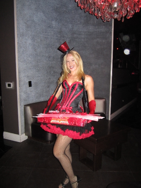 a woman dressed in a burlesquestyle costume stands in a nightclub.  she's wearing a red and black corset, a black tutu, fishnet stockings, red gloves, and a small top hat. she carries a tray of what appears to be candy. she is smiling and looks happy. the setting is dimly lit and appears to be upscale. the background includes a dark grey wall, a small table, and a chandelier partially visible in the upper left corner.