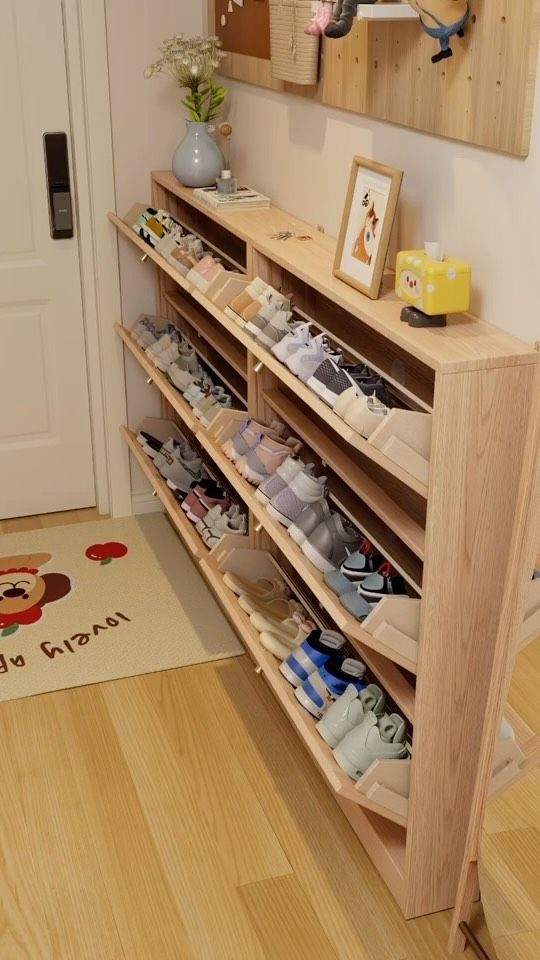 the image shows a wooden shoe rack filled with various pairs of shoes. it's a tilted design with multiple shelves. next to the shoe rack, there's a door and a rug with a cartoon design on the floor. on top of the shoe rack, there is a vase with flowers and a picture frame, along with other small decorative items. the shoe rack is made of lightcolored wood and seems to be part of the home decor, offering a storage solution for footwear.