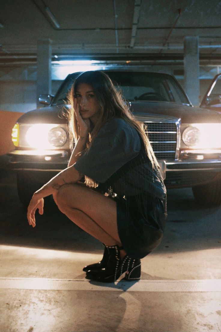 a young woman with long brown hair is squatting in front of a classic mercedesbenz car in a dimly lit parking garage. she's wearing a darkcolored short skirt, a gray tshirt, and black studded ankle boots. her expression is pensive as she rests her chin on her hand. the overall mood of the image is dark, moody, and stylish, highlighting the woman's fashion and attitude.  the car's headlights are visible, adding to the nighttime setting.