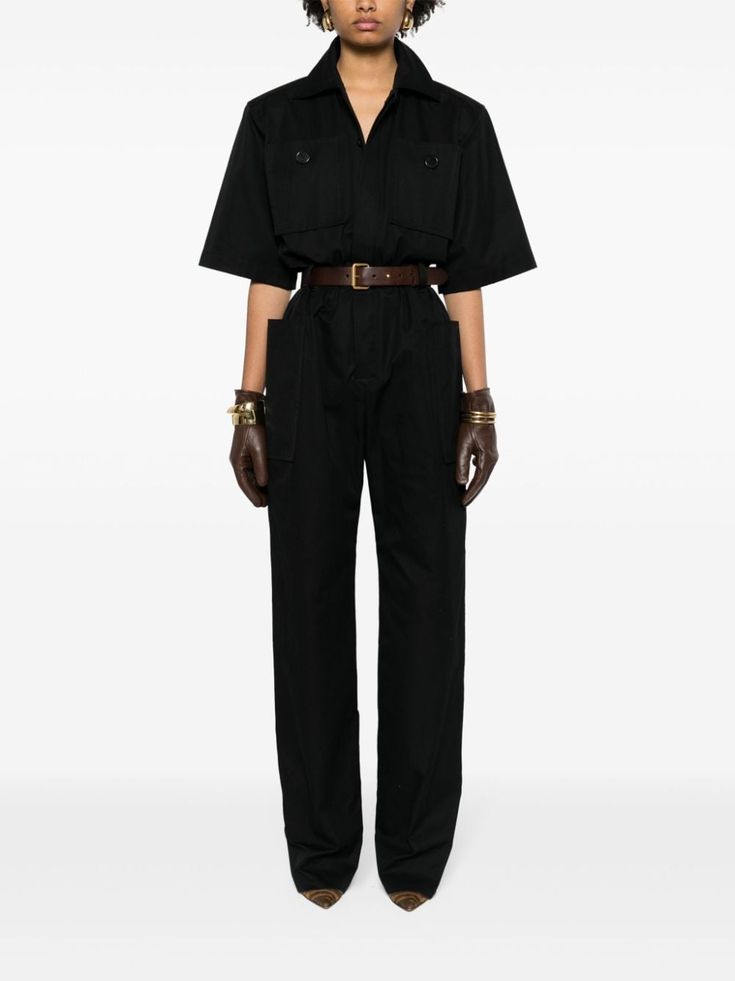 a woman wearing a black jumpsuit with brown belt and brown gloves, standing with a white background.