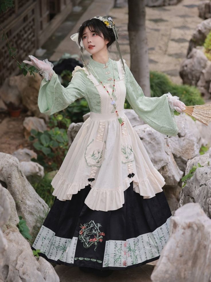 a woman in a beautiful, flowing hanfu dress stands in a garden, posing for the camera. the dress features a long black skirt with delicate floral embroidery and chinese calligraphy details, a creamcolored overskirt with lace trim, and a light green, longsleeved top with a high collar. her hair is styled in a traditional updo with decorative flowers. she holds a white lace fan in her left hand. the background features lush greenery, rocks, and a glimpse of a traditional chinese building. the lighting is natural and soft, creating a serene and elegant atmosphere.