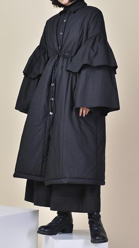 a woman wearing a long black coat with ruffled sleeves. the coat is buttoned up and has a cinched waist. she is wearing black boots. the background is a light beige. the woman is looking straight ahead with a serious expression.