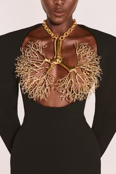 a woman in a black dress with long sleeves poses with her head tilted back. she is wearing a gold necklace that looks like branches.  the necklace is delicate and intricate. the woman has her hair pulled back, and her face is framed by a simple gold necklace. the background is a plain white studio wall.  the woman is looking directly at the camera.