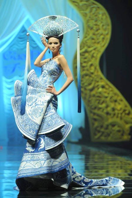 a woman models a blue and white porcelaininspired dress on a runway. she wears a matching headdress with tassels. the dress is strapless and features a layered skirt. the runway is reflective, and the backdrop is a mix of blue and gold.