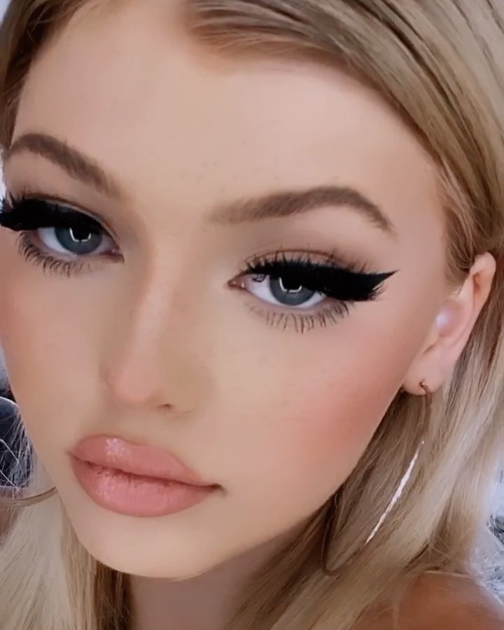 a closeup portrait of a woman with blonde hair, wearing heavy makeup. she has dramatic black eyeliner and false eyelashes, a nude lipstick, and blushed cheeks. she is wearing hoop earrings.