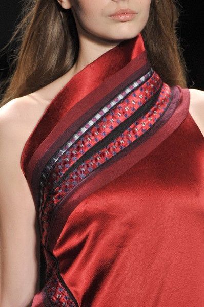 a closeup shot of a woman's red dress, featuring a unique collar detail. the collar consists of several layers of fabric with different patterns, creating a bold and eyecatching statement. the woman's long brunette hair cascades down her back, and her face is partially hidden in the collar, showcasing the intricate details of the garment. the overall look exudes elegance, sophistication, and high fashion.