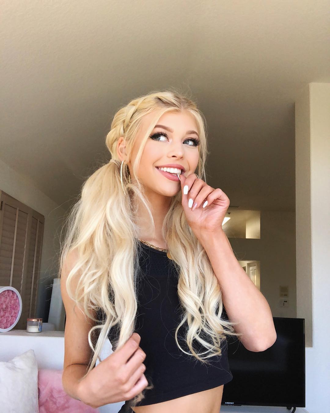 a woman with blonde hair in pigtails and braids is smiling and looking up. she is wearing a black tank top and gold hoop earrings. she has one finger in her mouth. the background shows an interior room with a tv, a shelf with decorations, and a couch with pink pillows.