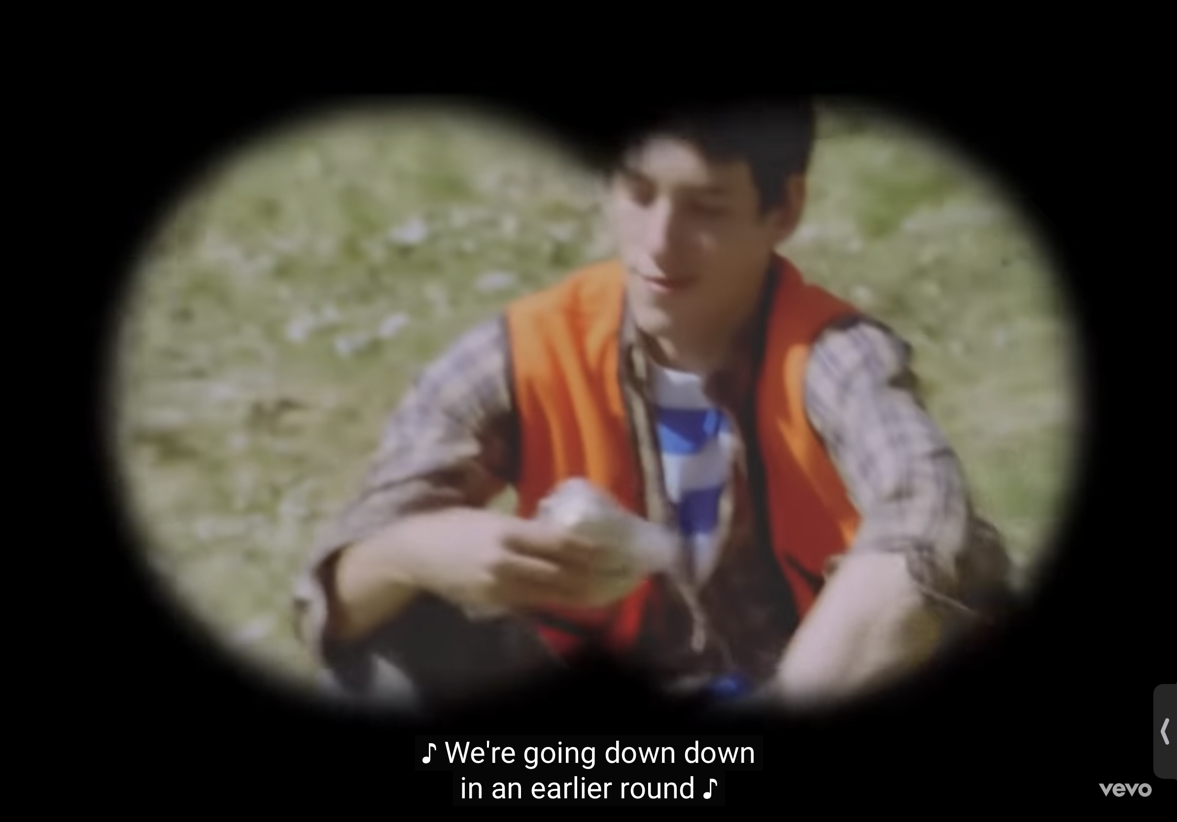 a man is sitting in a grassy field, looking through binoculars. he's wearing an orange vest and a white and blue striped shirt with a plaid jacket over the vest. he is holding a white object in his hands. the binoculars are focused on him, making the image appear circular. the image is taken from the point of view of someone looking through the binoculars.