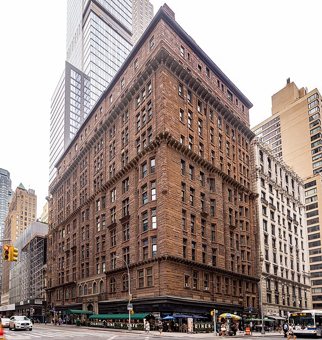 a photo of a brownstone building that takes up the majority of the frame. there are cars and buses driving on the street in front of it. there are storefronts on the ground floor of the building. there is a taller, more modern glass skyscraper behind the brownstone building. there are other buildings in the background on either side of the brownstone building. the sky is overcast.