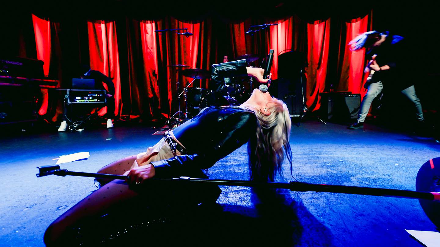 a female singer performs on stage during a rock concert. she is kneeling and leaning back, singing into a microphone. a microphone stand is in front of her. she is wearing a black outfit and has long blonde hair. behind her, the stage is set with various musical instruments including drums, a keyboard, and a guitar. other band members are present, and red curtains hang in the background. the stage is illuminated with blue and red lighting, creating a dramatic atmosphere.