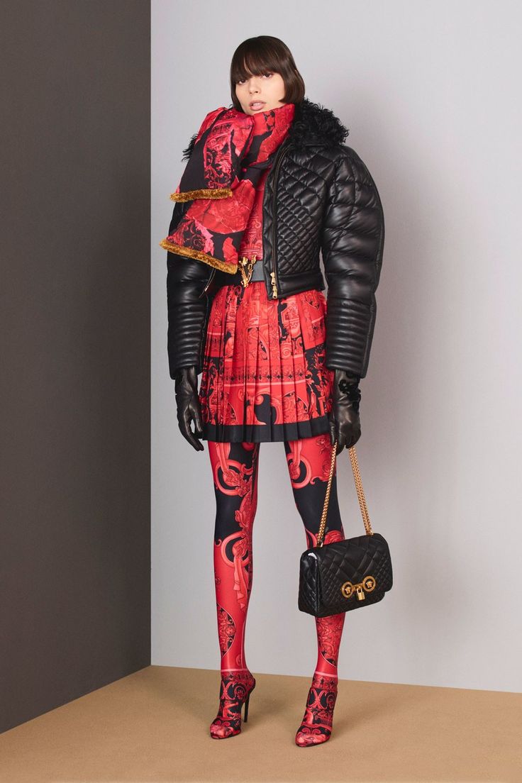 a full shot of a fashion model showcasing a versace outfit. the model, with short brown hair, is wearing a black quilted leather jacket with a large collar, a red pleated skirt with a black and red pattern, matching patterned tights, and black leather gloves. she has a red and black patterned scarf around her neck. she's also carrying a black versace handbag with a gold clasp and wearing heeled mules that match her tights. the background is a simple gray and dark gray wall.