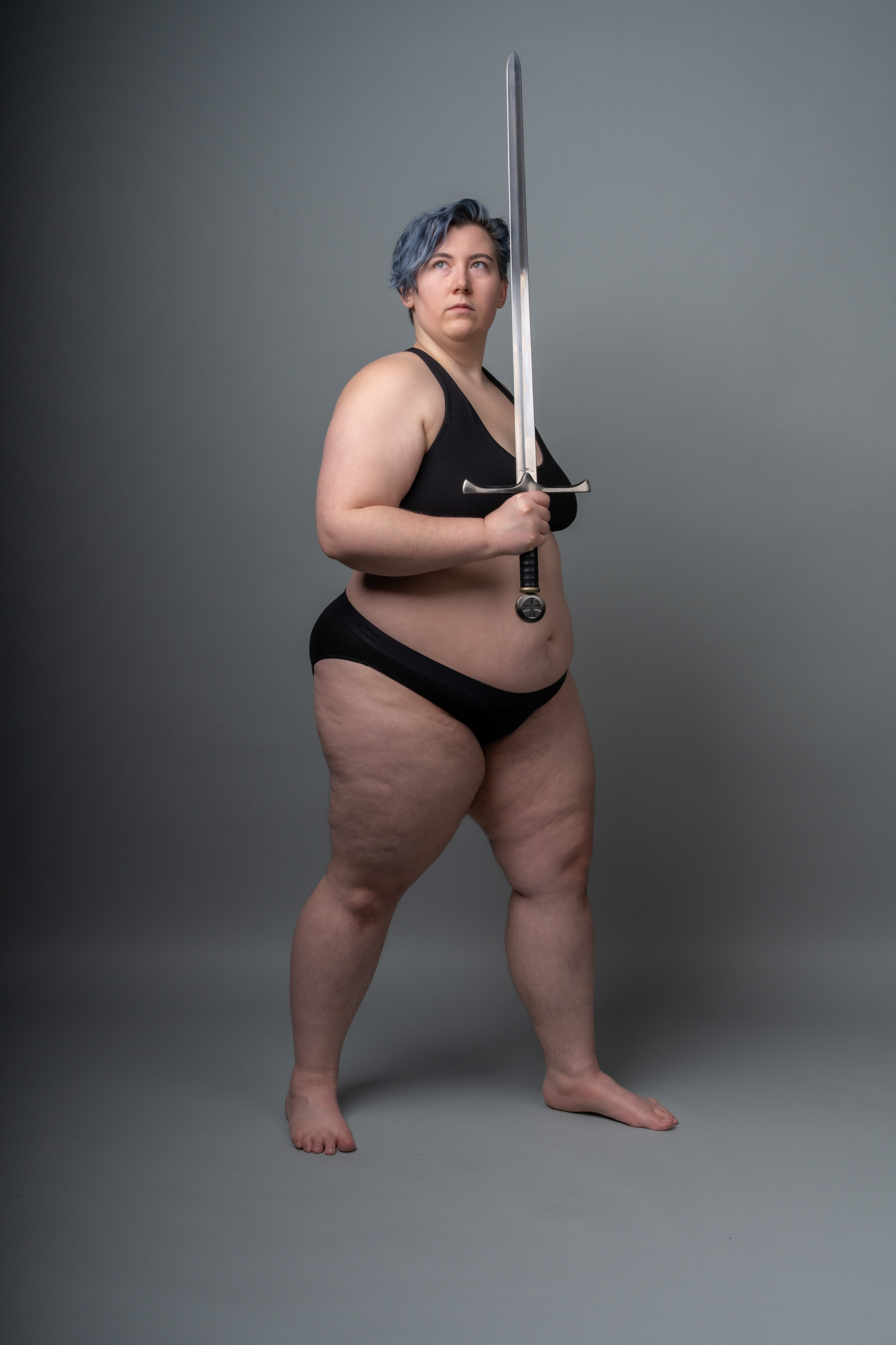 a full shot of a plus size woman with short blue hair holding a sword up high. she is wearing a black bra and black bikini bottoms, and she is standing in a studio with a grey background. she is looking up.
