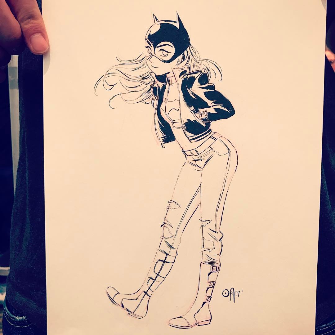 a black and white drawing depicts batgirl in a fulllength pose. she is wearing her signature bateared mask, a jacket, jeans, and boots. the artwork is held up, partially obscuring the edges with hands.