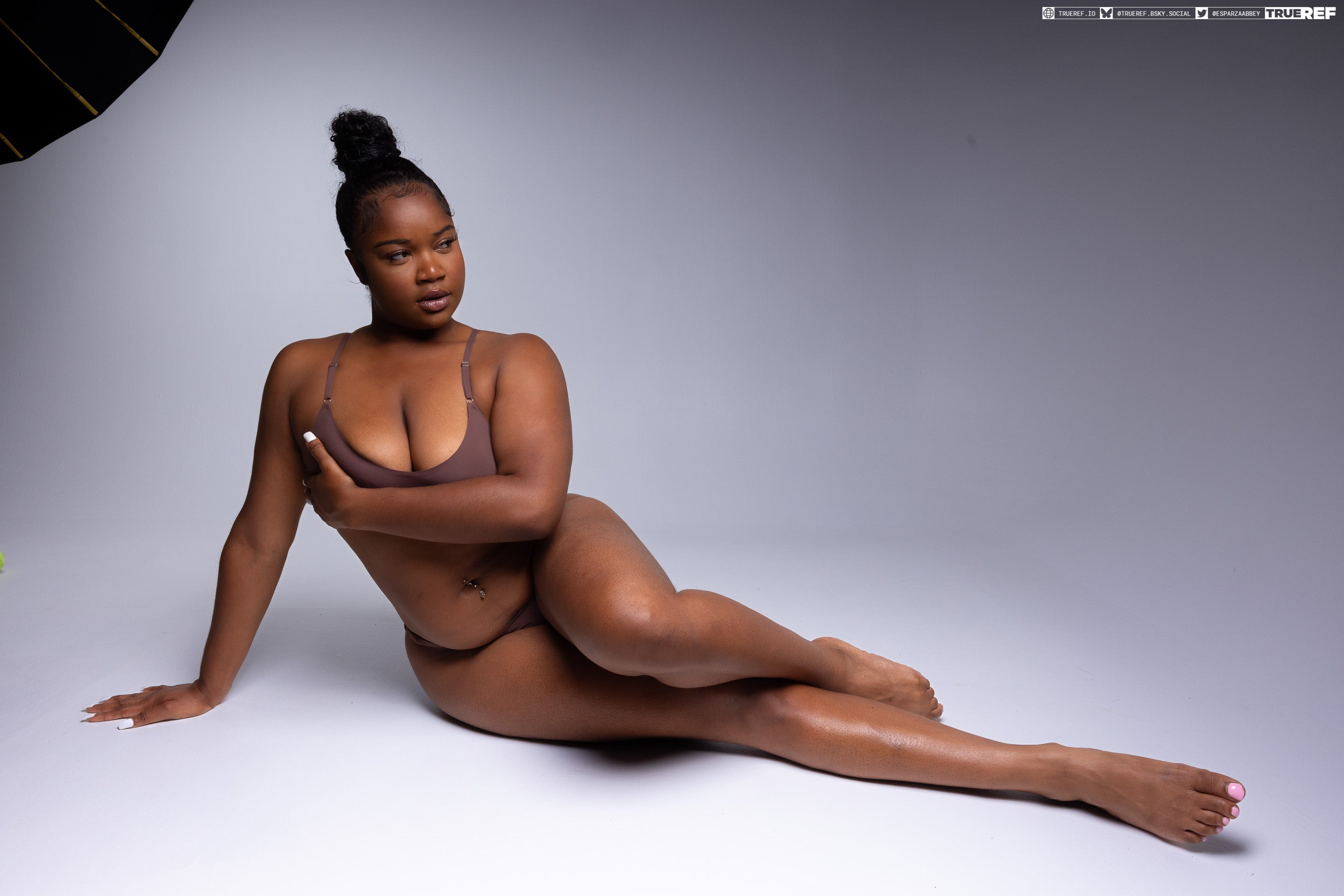 a beautiful darkskinned woman is lying down in a studio shot. she is wearing brown lingerie and has a confident and attractive pose. her hair is up, and she has a belly button piercing. the background is light. she has white nails and dark eyelashes. she is looking away from the camera. the image shows her bare shoulders and legs. the lighting is soft and flattering.