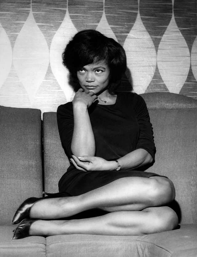 a black and white portrait of a woman sitting on a couch. she is wearing a black dress and has her hair styled in a short, bouffant style. she is looking away from the camera, and she has a thoughtful expression on her face. the photo has a vintage feel and was likely taken in the 1960s.