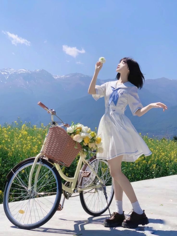 a young asian woman in a white sailor collar dress stands next to a pastel yellow cruiser bike with a basket of yellow flowers. she is holding a single flower in her hand and looks up towards the bright blue sky, with snowcapped mountains in the background.  the scene is idyllic and cheerful, showcasing a bright sunny day in a picturesque natural setting. the overall aesthetic is light, airy, and peaceful.
