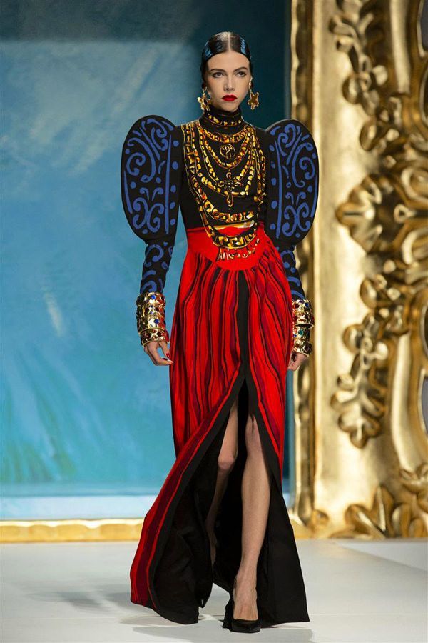 a fashion model poses on a runway wearing an ornate gown. the gown features puffed sleeves decorated with blue designs on a black background, a black turtleneck bodice with multiple gold necklaces, and a highwaisted red skirt with black lining and a high slit revealing the model's leg. the model is also wearing large gold earrings and multiple gold bracelets on each wrist. the background includes a blue painted backdrop and a baroquestyle golden frame.