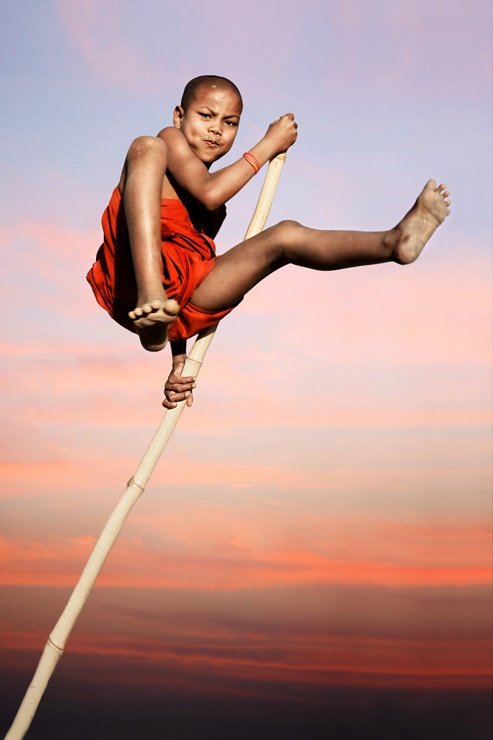 a young boy in a red robe is jumping on a bamboo pole against a backdrop of a stunning sunset sky. he is smiling widely, his legs extended, and he is airborne. the image captures the joy and energy of youth. the vivid colors of the sunset provide a beautiful contrast to the boy's silhouette.