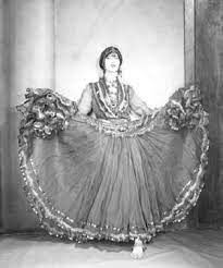 a black and white fulllength shot depicts a woman wearing a traditional dress, likely for a performance or dance. the woman is adorned in a decorative headdress and an elaborate skirt with ruffled edges, fully extended to display its wide circumference. she stands in front of what appears to be a stage backdrop or set design. the image has a vintage aesthetic, reminiscent of older photography styles.