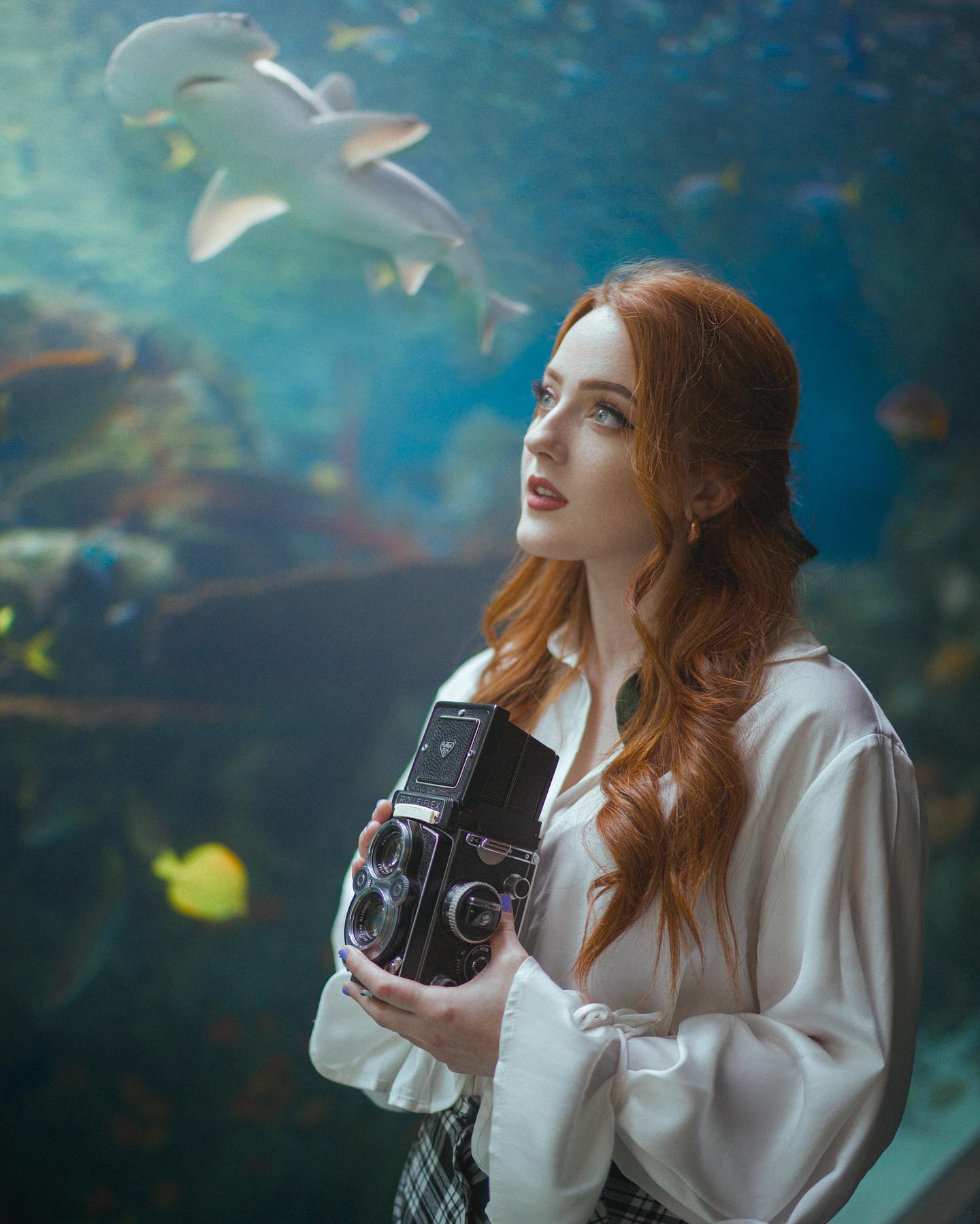 a young woman with long red hair stands in front of a large aquarium tank. she is holding a vintage camera in her hands and is looking up at a shark swimming above her. the aquarium is lit with artificial light, creating a cool and serene atmosphere.
