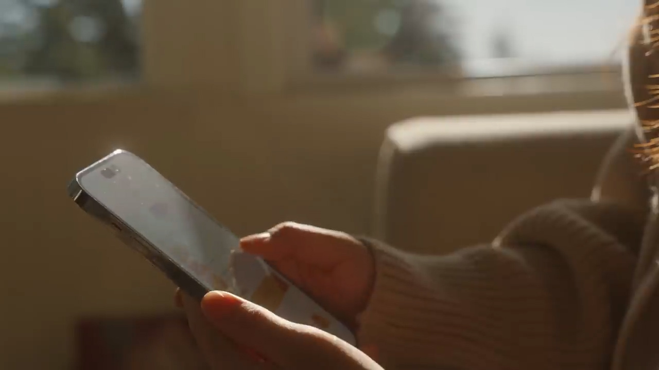 a closeup shot of a woman's hands holding a smartphone. the woman is wearing a beige sweater and sitting in a sunlit room. the image has a warm and cozy feel.
