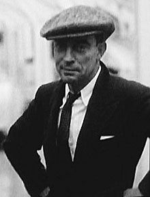 a black and white medium shot of a man wearing a suit, tie, and newsboy cap standing outside. he has his hands on his hips.
