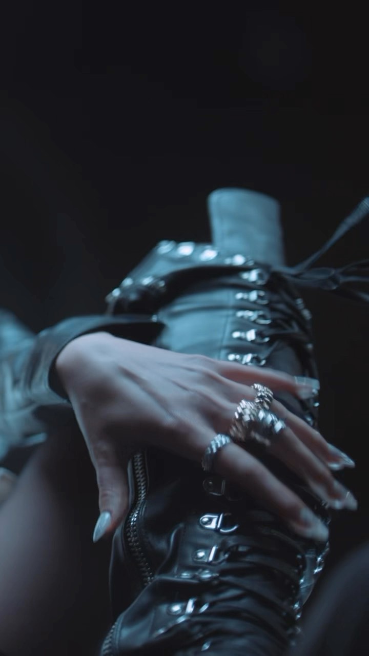 a closeup shot captures a hand adorned with several silver rings resting on a black leather, laceup boot. the hand features long, elegant nails, possibly enhanced with nail art. the boot is detailed with metal hardware and laces, showcasing a stylish, edgy fashion aesthetic. the dark background emphasizes the textures and details of the jewelry and leather.
