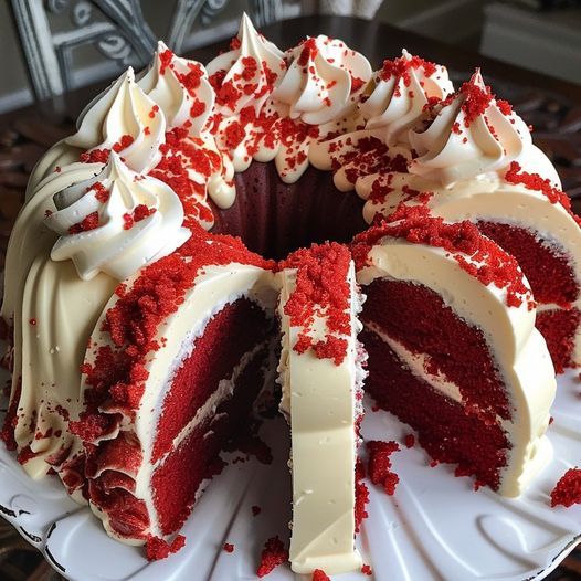 a closeup shot of a beautifully decorated red velvet bundt cake.  the cake is frosted with a generous layer of cream cheese frosting and topped with red velvet sprinkles. several slices have been cut, revealing the moist, deep red layers inside. the cake sits on a white cake stand, enhancing its presentation. the overall image is highly appetizing, emphasizing the cake's texture and richness.