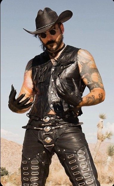 a man in a black cowboy hat, black sunglasses, and a black leather jacket stands in a desert. he is wearing leather pants and a belt with silver buckles. he has a long beard and tattoos on his arm. he is looking at the camera.