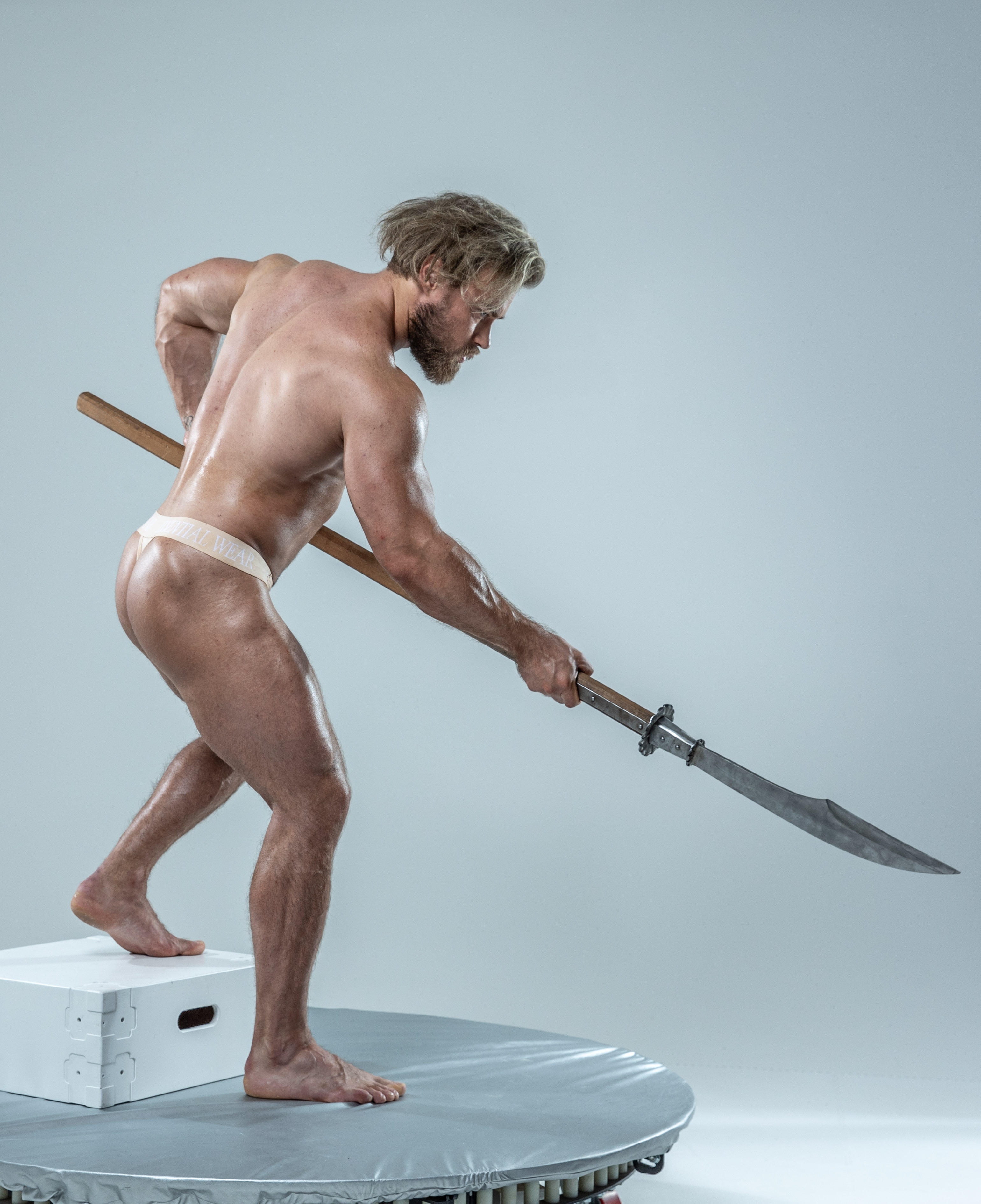the image showcases a muscular man in a dynamic pose, wielding a spearlike weapon. he stands on a white platform against a seamless gray backdrop. the soft lighting accentuates his physique and the spear's metallic sheen.  his focused expression and poised stance convey a sense of strength and readiness. the overall aesthetic evokes themes of ancient warriors or mythological figures.