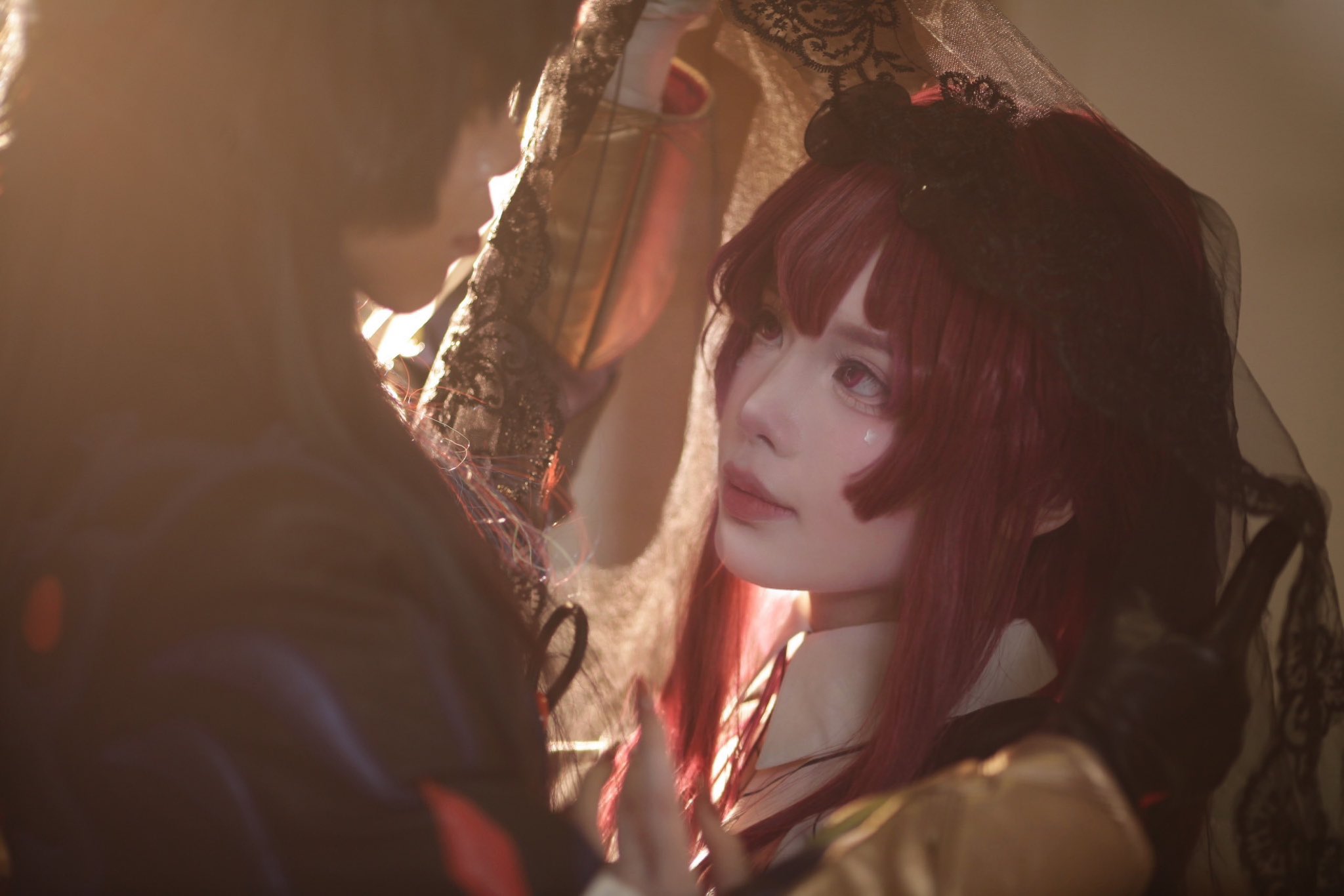 a portrait of a woman with long red hair, dressed in a black dress and veil. she looks at the camera with a gentle and mysterious expression. she's holding the veil with her right hand, and her left hand is resting on her chest. the lighting in the photo is soft and warm, creating a romantic atmosphere.
