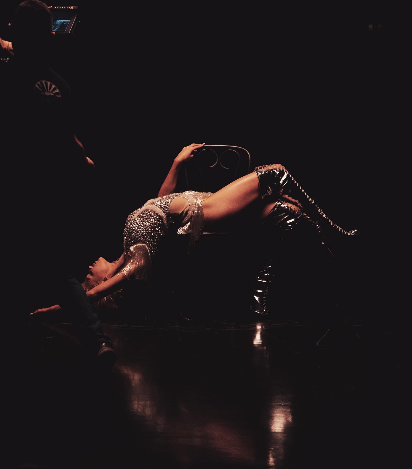 the image shows beyonce lying down in a backbend position, her upper back resting on a chair with her knees bent and wearing highheeled boots. she is facing away from the camera and wearing a sparkly outfit. the background is dark, suggesting the image was taken during a stage performance. there is a person standing to the left of beyonce and some light is reflecting off the stage floor.
