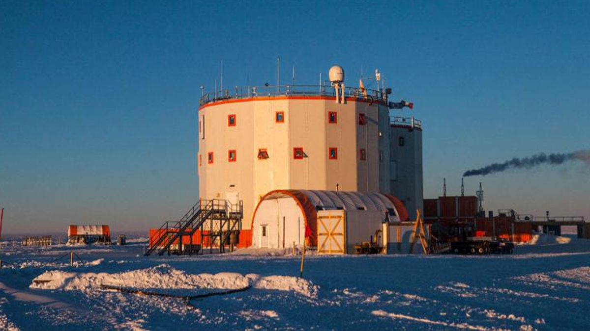 a large, white research station sits on a snowy, icy landscape. the station appears to be made of several interconnected buildings, with a large central tower. there is smoke coming from a chimney on the left side of the station, indicating that it is in operation. the surrounding landscape is covered in snow and ice, and the sky is a clear blue. this image captures the harsh yet beautiful environment of the arctic or antarctic, where scientists conduct research on a variety of topics.
