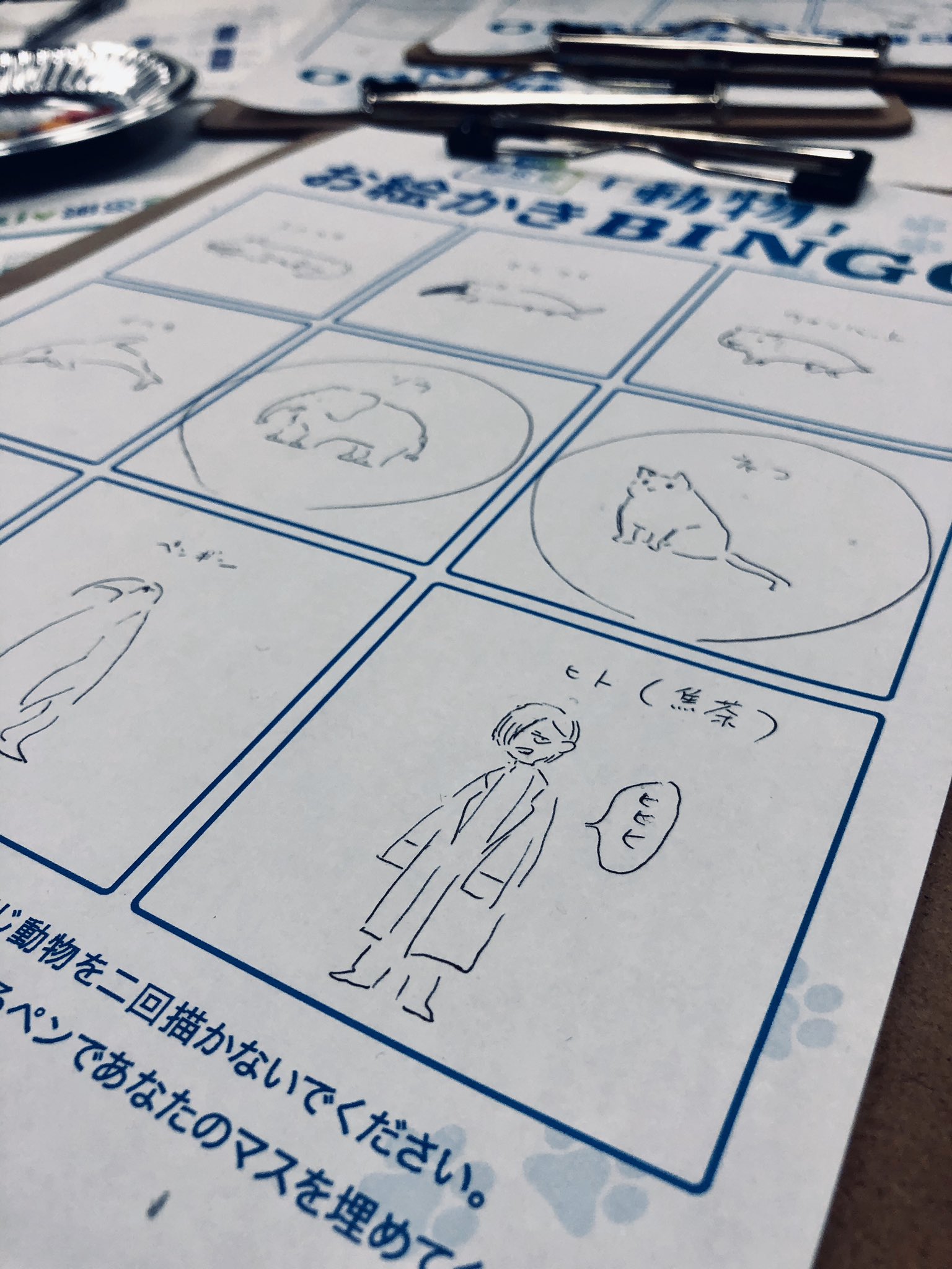 a closeup image of a bingo card drawn on a piece of white paper. the bingo card features simple line drawings of various animals, including a dolphin, penguins, a cat, and a person. the text on the card is written in japanese. there is a black pen clipped to the top of the paper.