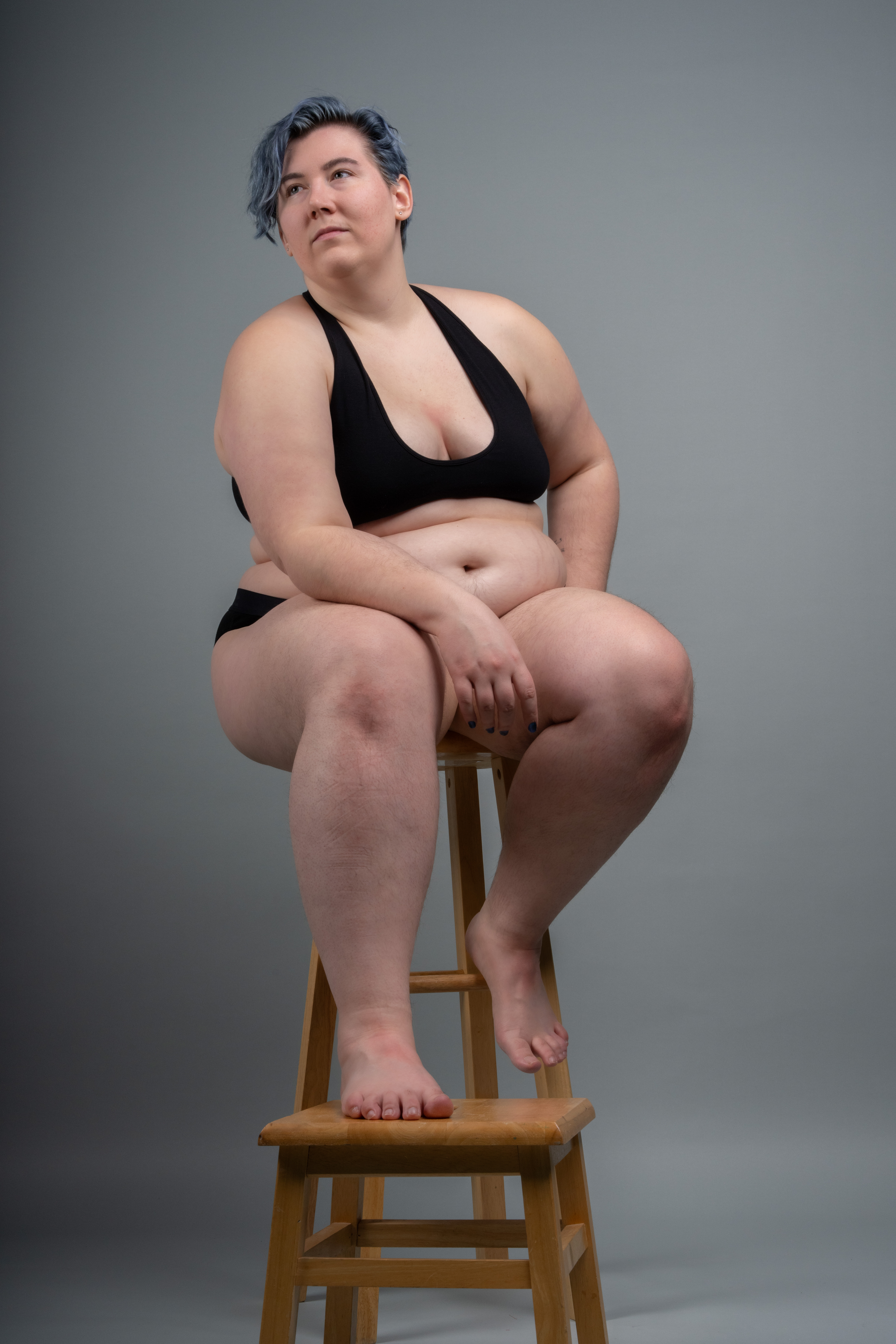 a plussize woman with blue hair sits on a wooden stool in a studio setting. she is wearing a black bra and black underwear. the background is gray. she is looking up and away from the camera.