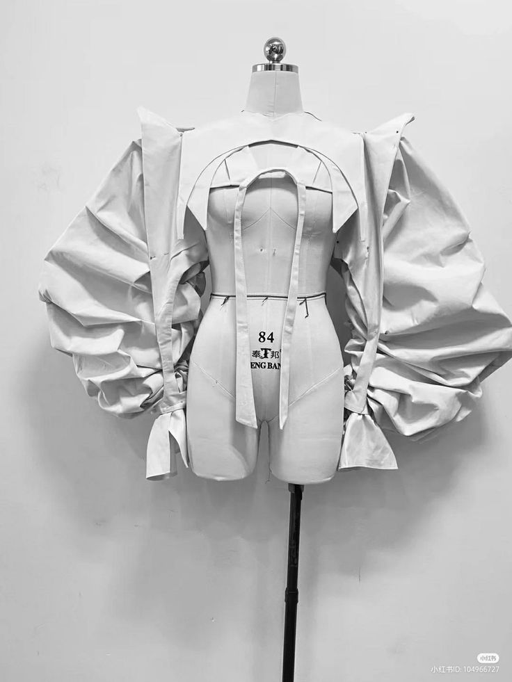 a white dress form is wearing a white fabric garment that is draped over it. the fabric is bunched up in a way that would be difficult to sew and the design is almost futuristic. the dress form is in a studio setting, and is ready for the garment to be sewn. the dress form is a popular tool for fashion designers and dressmakers, and is used to create and fit garments. the dress form is a timeless tool that is used to create beautiful and functional garments. the dress form is also a great way to learn about fashion design and the construction of clothing.