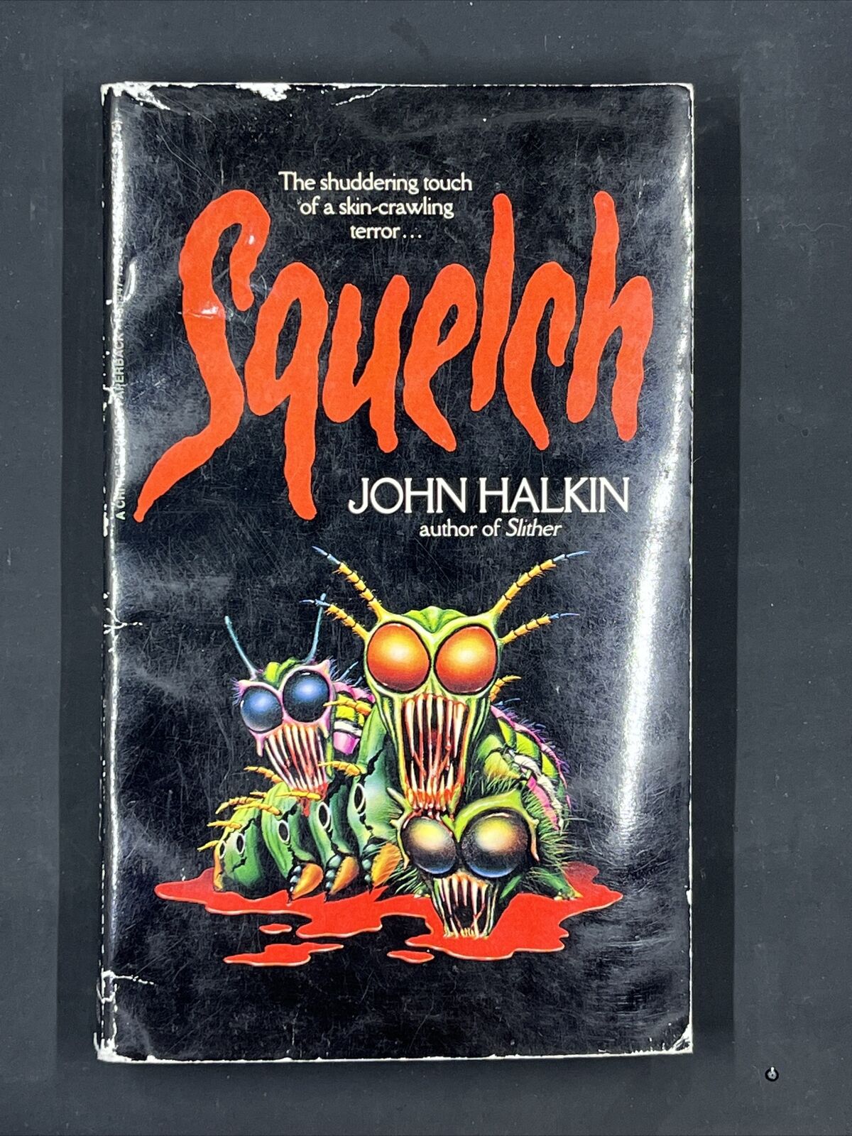 loseup view of a vintage paperback book titled "squelch" by john halkin. the cover features a striking illustration of several grotesque, insectlike creatures with sharp teeth and glowing eyes, surrounded by blood. the title is written in large, stylized red letters, with the author's name and a tagline ("the shuddering touch of a skincrawling terror...") underneath. the book's overall aesthetic suggests a horror or science fiction theme, typical of paperback novels from the 1980s.  the book shows some signs of wear consistent with age.